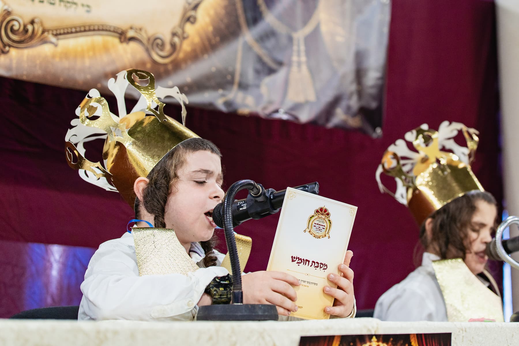 A boy holding a Mesibat Chumash booklet and reciting into a microphone
